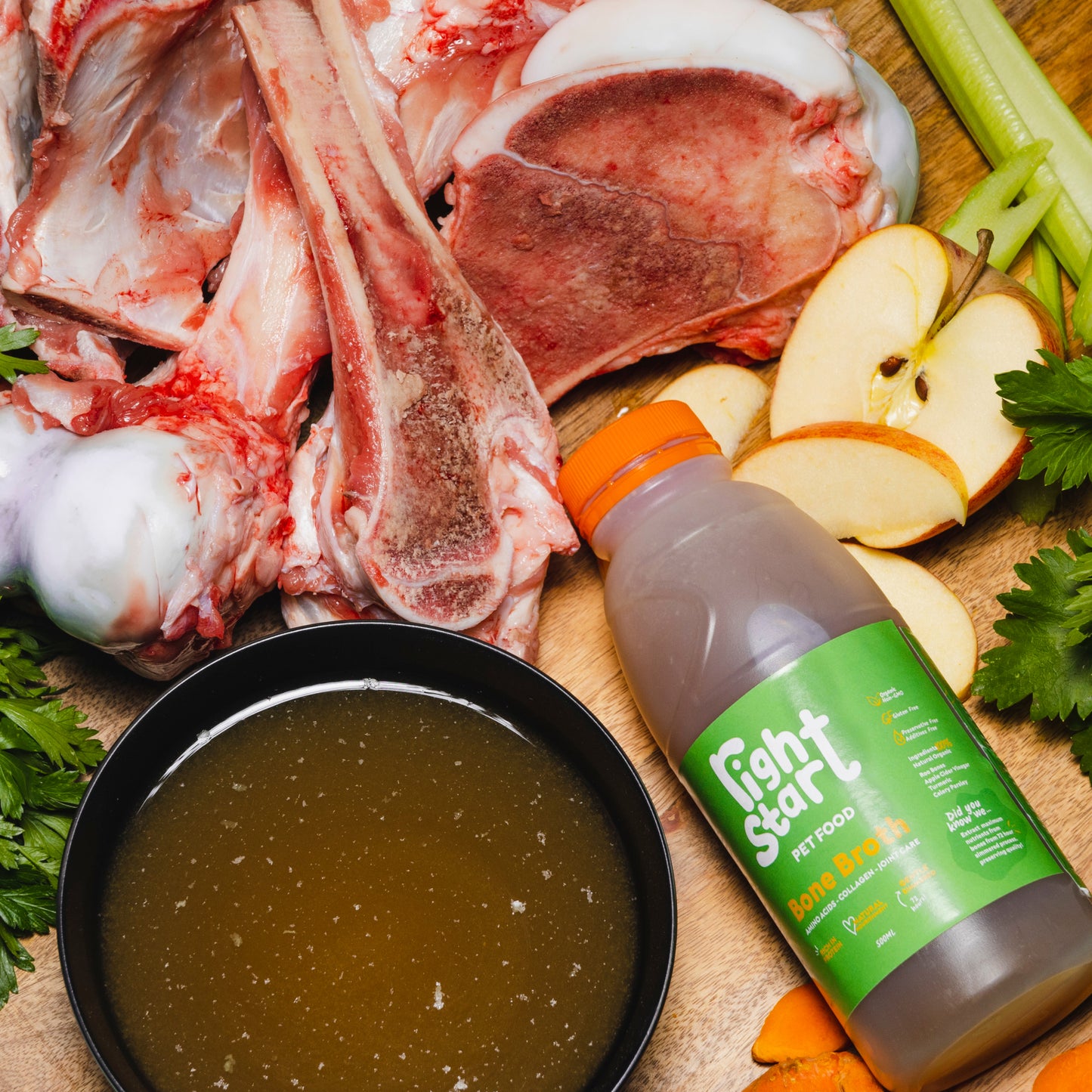 right start pet food bone broth. broth bottle on right, black bowl with broth on left, raw bones in background, apple and celery